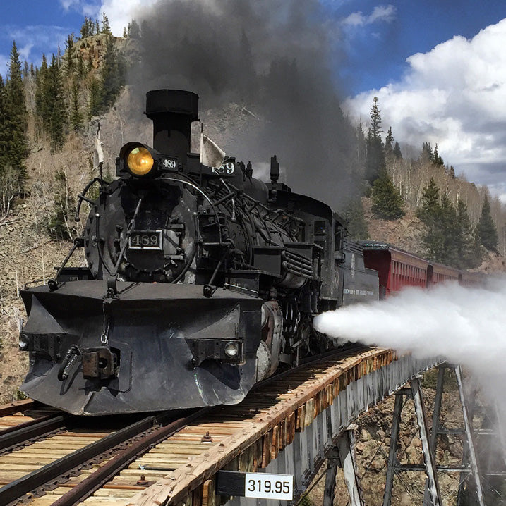 Steam Train going across raised wooden bridge with mountain in background. The bridge has a white sign with black numbers 319.95.
